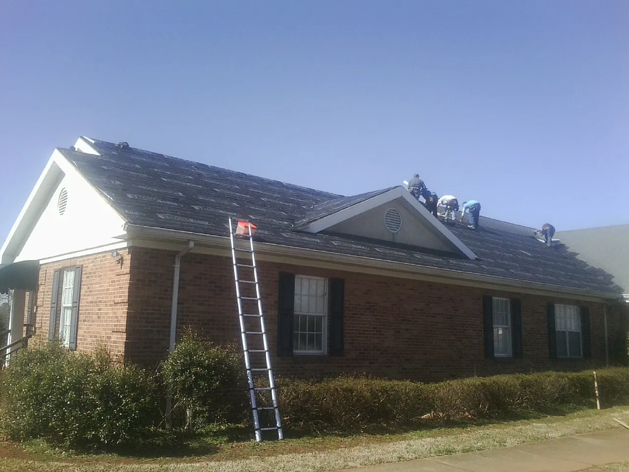 Crew tearing shingles off a brick ranch roof with a ladder set — Emergency Roof Repair work by our crew in Conetoe