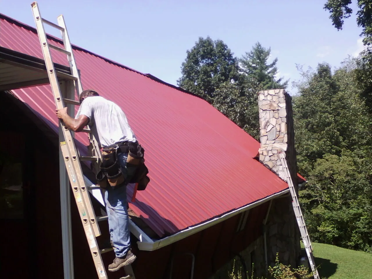 Roofer on a ladder beside a red metal roof and stone chimney — Soffit & Fascia Repair by Roofing Brothers in Conetoe