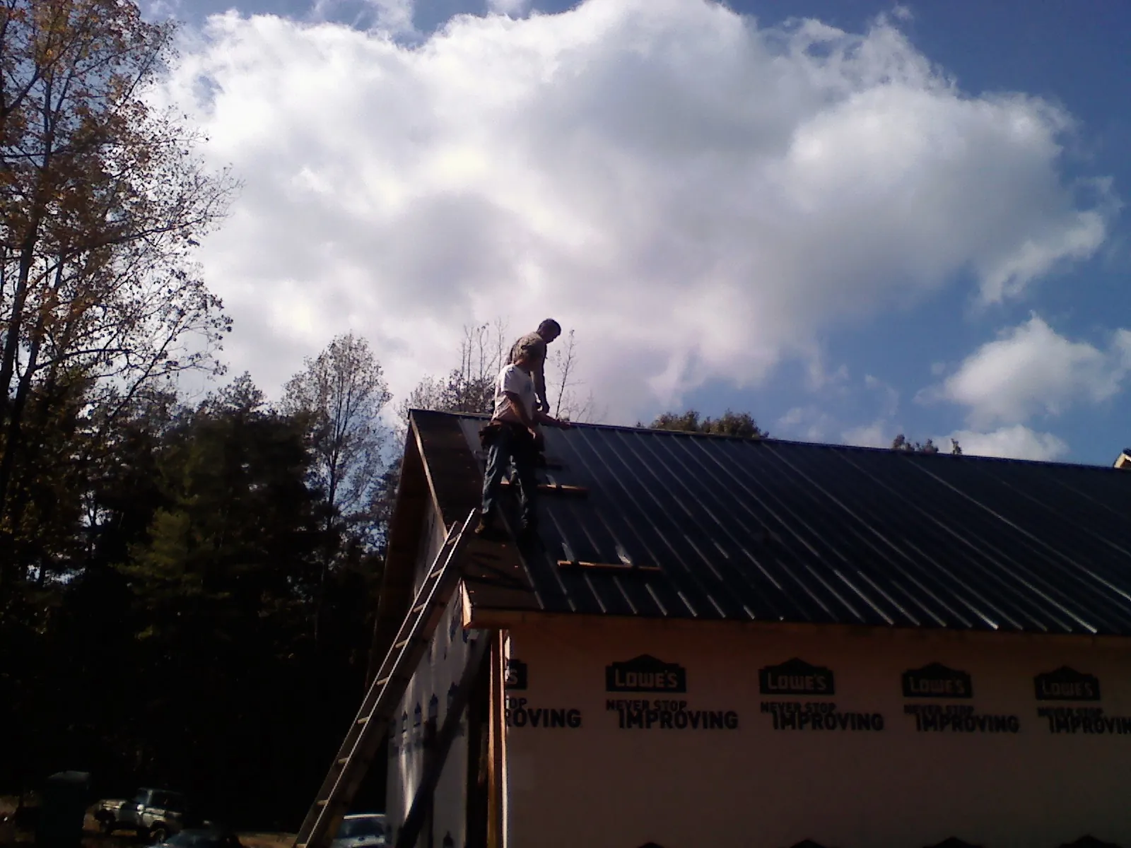 Roofer fastening dark metal panels high against a cloudy sky, from a Roof Inspection job in Conetoe